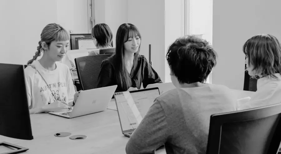 4人の男女が同じデスクでPCを開きながら会議をしている様子